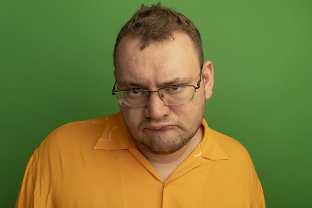 皱眉戴着眼镜和橘色衬衫的男人不满地皱着眉头站在绿色的墙上眼镜男人站着