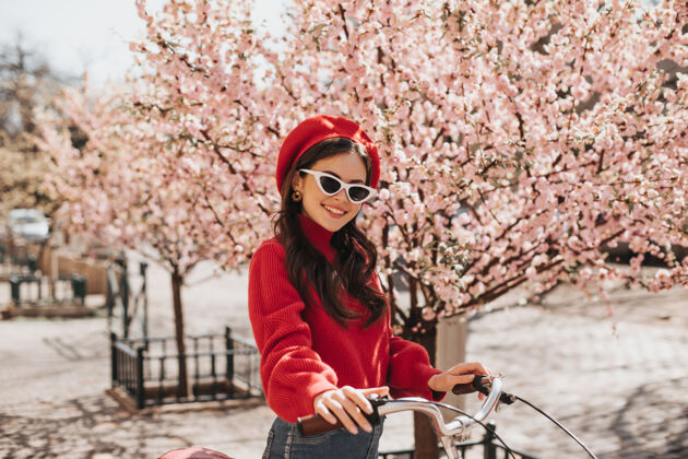春天黑发女孩戴着红帽子和毛衣在樱花的背景下摆姿势迷人的女人戴着时髦的太阳镜微笑着骑着自行车女性贝雷帽温柔