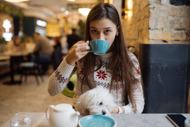 杯子美丽的女人抱着她的可爱的狗 喝着咖啡 微笑着在咖啡馆人年轻餐馆