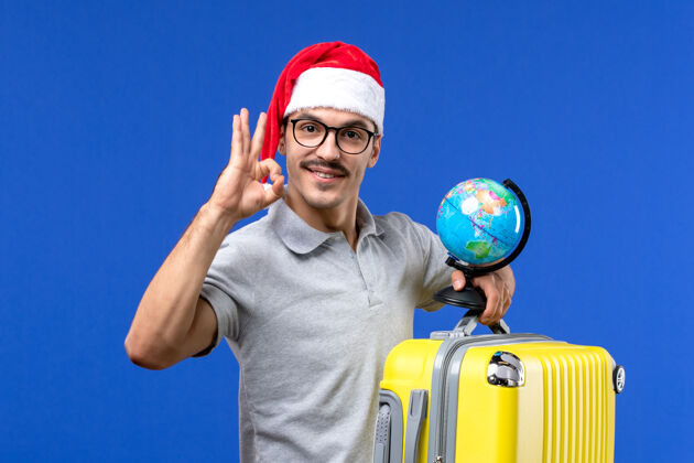 运动正面图年轻男子手持地球仪和黄包上蓝墙飞机度假之旅举行表演浴帽