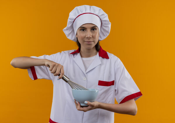 女孩穿着厨师制服的年轻快乐的白人厨师女孩拿着威士忌和碗孤立地放在橙色的空间里厨师复制搅拌