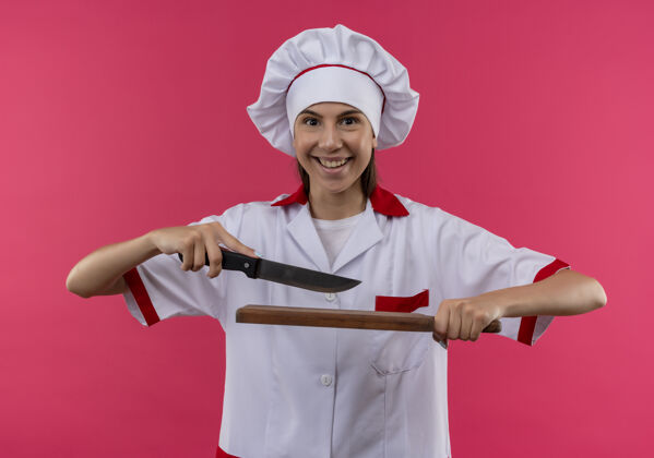 烹饪年轻惊讶的白人厨师女孩穿着厨师制服拿着刀和砧板隔离在粉色空间和复印空间持有女孩粉红