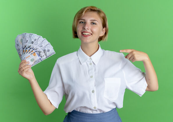 俄罗斯年轻的金发碧眼的俄罗斯女孩抱着钱 指着隔离在绿地上的复制空间持有年轻金钱