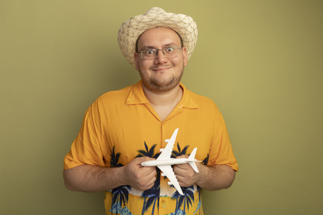 正面戴着眼镜的男人穿着橘色衬衫戴着夏天的帽子拿着玩具飞机快乐而积极的微笑站在绿色的墙上眼镜夏天微笑