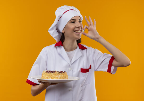 蛋糕身着厨师制服的年轻微笑的白人厨师女孩拿着盘子上的蛋糕 在橙色的空间和复制空间上做着美味的手势年轻标志空间