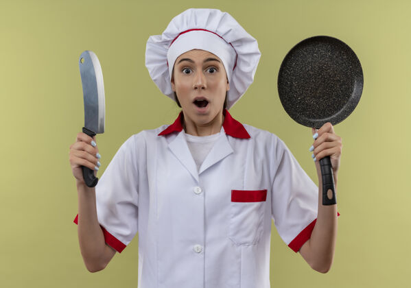 女孩穿着厨师制服的年轻白人厨师女孩拿着刀和煎锅孤立地放在绿色空间里厨师复制绿色