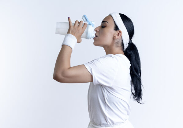 立场年轻自信的漂亮运动女人戴着头带和腕带站在一边喝水隔离在白色空间与复制空间侧空间年轻
