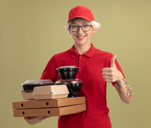制服年轻的送货员身穿红色制服 戴着帽子 戴着眼镜 手里拿着披萨盒和食品包 面带微笑 站在绿色的墙上竖起大拇指披萨帽子脸