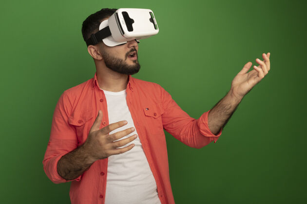 胡须身穿橙色衬衫 戴着虚拟现实眼镜 留着胡子的年轻人站在绿色的墙上做手势虚拟站立年轻