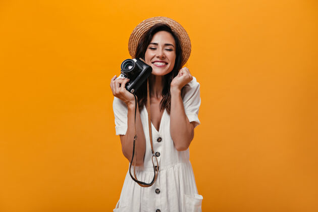 室内心情愉快的女人拿着复古相机在橙色的背景下漂亮的女孩在轻时尚的衣服和草帽摆姿势连衣裙帽子休闲