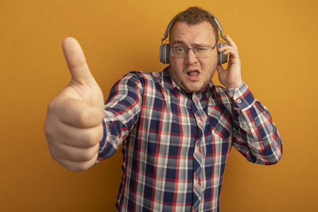 检查一个戴着眼镜和格子衬衫 戴着耳机的男人站在橙色的墙上 自信地竖起大拇指耳机男人自信