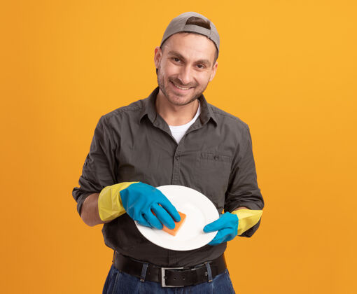举行年轻的保洁员穿着休闲服 戴着橡胶手套 拿着盘子和海绵 站在橘色的墙上 满脸笑容 自信满满清洁年轻橡胶