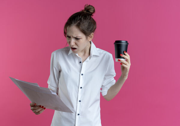 年轻年轻恼怒的金发俄罗斯女孩看着纸页上拿着咖啡杯孤立的粉红色空间与复制空间杯子看空间