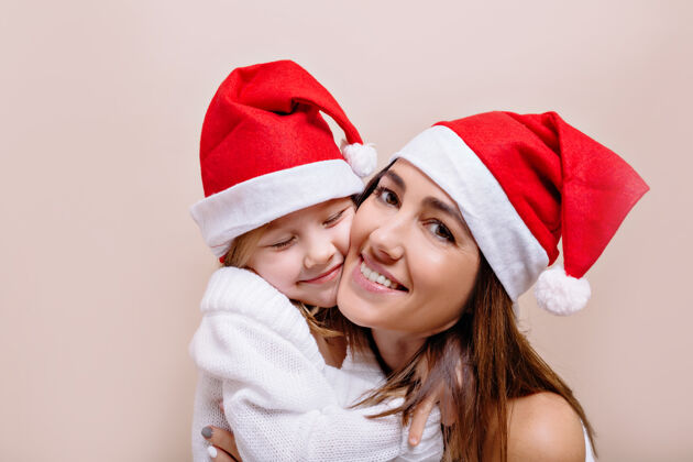 圣诞快乐 微笑 有趣的母女俩戴着圣诞老人的帽子摆姿势 抱着脸一个年轻漂亮的女人 嘴唇明亮 抱着一个5岁的女孩女人孩子圣诞帽