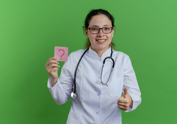 年轻快乐的年轻女医生 穿着医用长袍 戴着听诊器 戴着眼镜 拿着问话单 大拇指竖起 隔离在绿色的墙上 留着复印空间穿拇指看