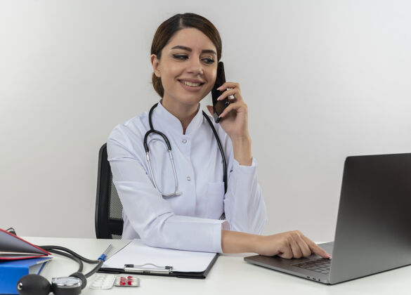 长袍面带微笑的年轻女医生穿着医用长袍和听诊器坐在办公桌旁 手里拿着医疗工具 看着笔记本电脑 隔着白墙上的电话聊天工具看办公桌