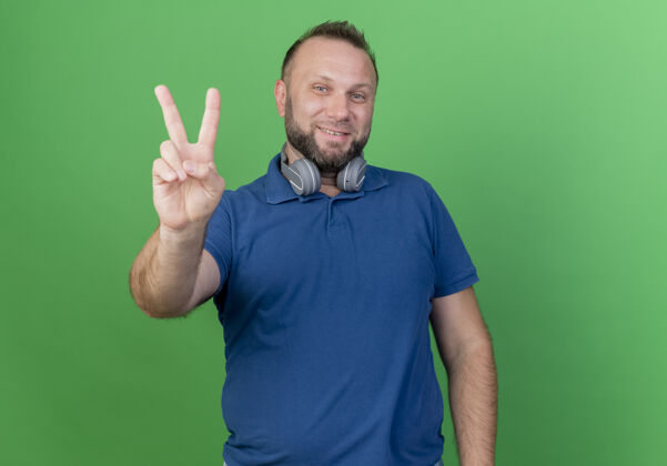 男人微笑的成年斯拉夫男子戴着耳机在脖子上看做和平标志隔离在绿色墙壁与复制空间穿微笑斯拉夫