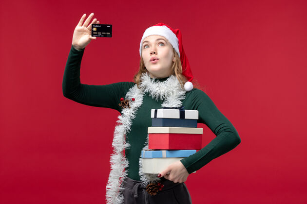 姿势正面图：手持礼物和红色背景的银行卡的年轻女子银行视图圣诞节