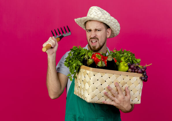斯拉夫年轻英俊的斯拉夫园丁穿着制服 戴着帽子 手里拿着一篮子蔬菜和耙子 孤零零地站在深红色的墙上制服耙子篮子