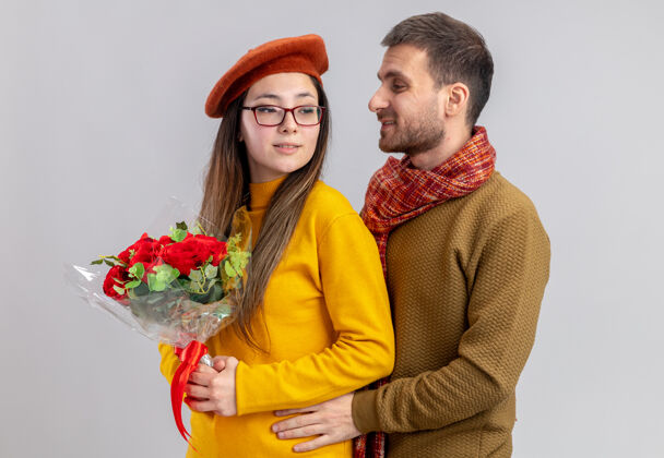 日子年轻漂亮的情侣幸福的男人抱着他微笑的女人 捧着一束红玫瑰 一起欢庆情人节 站在白墙上花束微笑女人