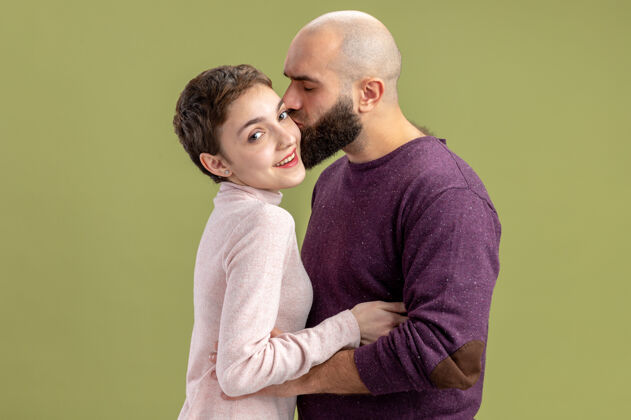 立场一对穿着休闲装的年轻夫妇一个留着短头发和胡须的女人一个热恋中的男人站在绿色的墙上亲吻他的女友庆祝情人节休闲白天胡须