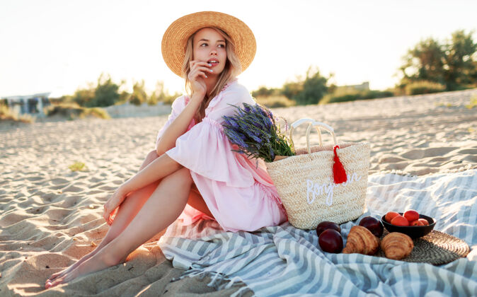 户外在海边的乡间野餐优雅的年轻女子 金色卷发 穿着优雅的粉色连衣裙 享受假期 吃水果微笑女性休息