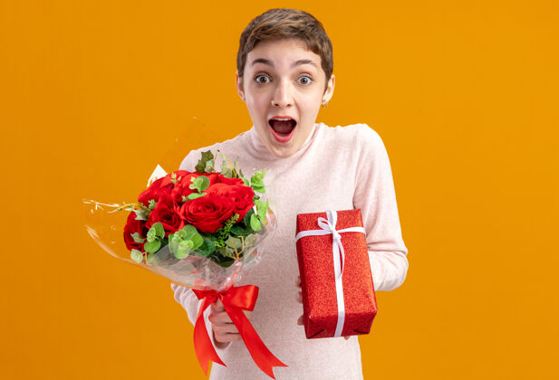 年轻一位留着短发的年轻女子手持一束红玫瑰和一件礼物 看着相机 站在橙色的墙上 惊讶地看着情人节的概念天举行玫瑰