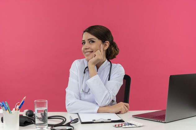 放面带微笑的年轻女医生穿着医用长袍和听诊器坐在办公桌旁 手里拿着医疗工具和笔记本电脑 双手放在办公桌上 下巴看起来笔直地孤立在粉红色的墙上微笑医疗手