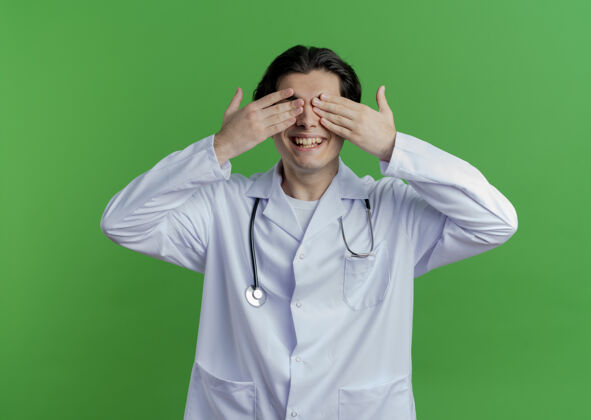 长袍面带微笑的年轻男医生穿着医用长袍和听诊器 双手隔离在绿墙上 遮住眼睛微笑盖穿