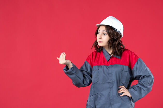 微笑特写镜头中惊讶的女建筑工人穿着制服 戴着安全帽 在隔离的红墙上呼唤着某人有人成功人