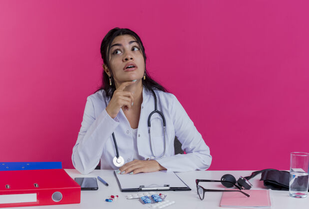 听诊器年轻的女医生穿着医用长袍 手持听诊器坐在办公桌旁 手持医疗工具 手放在办公桌上 一边看着一边 一边抚摸着粉红色墙上的下巴放工具穿