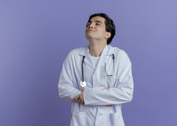 长袍高兴的年轻男医生穿着医用长袍和听诊器 手放在肚子上 另一只手放在胳膊上 闭着眼睛 隔离在紫色的墙上 留有复印空间另一个封闭医疗