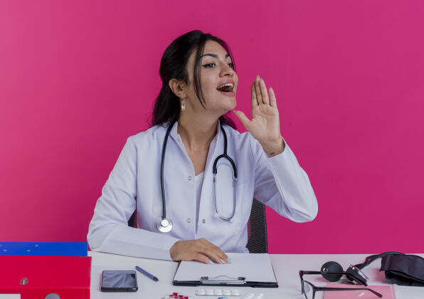 空气年轻的女医生穿着医用长袍 手持听诊器坐在办公桌旁 手持医疗工具 手放在办公桌上 一边看着一边 手举在空中 对着隔离在粉色墙壁上的人喊叫听诊器手长袍