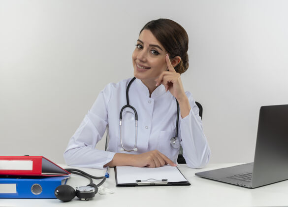 笔记本电脑面带微笑的年轻女医生穿着医用长袍和听诊器坐在办公桌旁 手里拿着医疗工具和笔记本电脑 手指被隔离在白墙上 触摸着头部穿头听诊器