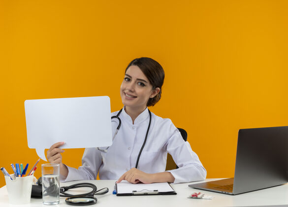 医生面带微笑的年轻女医生穿着医用长袍和听诊器坐在桌子旁 手里拿着医疗工具和笔记本电脑 手里拿着隔离在黄色墙上的聊天泡泡笔记本电脑工具长袍