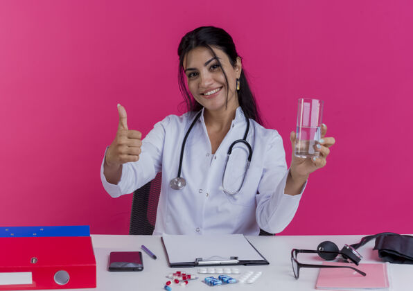 坐着微笑着的年轻女医生穿着医用长袍和听诊器坐在办公桌旁 手里拿着医疗工具 手里拿着一杯水 在粉红色的墙上孤立地竖起大拇指表演微笑长袍