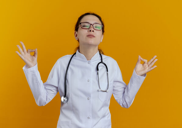 穿着年轻的女医生闭着眼睛 穿着医用长袍 戴着听诊器 戴着眼镜 在橘黄色的墙上显示出冥想的隔离橙色长袍表演