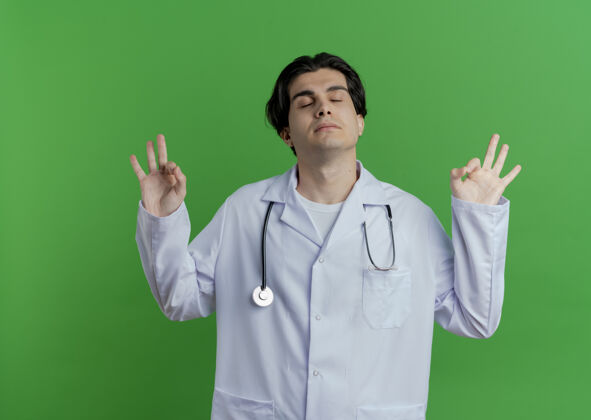 封闭和平的年轻男性医生穿着医用长袍和听诊器 闭着眼睛做着正常的迹象 隔离在绿色墙壁上 留有复印空间好的年轻听诊器