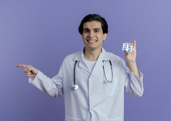 指着面带微笑的年轻男医生 穿着医用长袍 手持听诊器 指着隔离在紫色墙上的一侧的一包胶囊长袍年轻胶囊