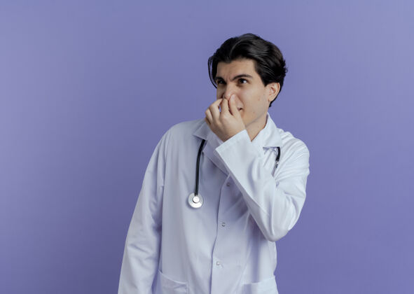 长袍恼怒的年轻男医生穿着医用长袍 手持听诊器 鼻子望着隔离在紫色墙上的一边 留着复印空间壁板医疗鼻子