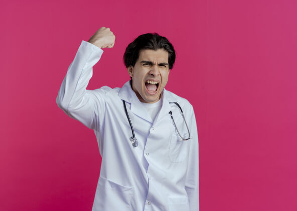 长袍快乐的年轻男医生穿着医用长袍和听诊器做着“是”的手势 隔离在粉红色的墙上 留着复印空间穿着表情听诊器