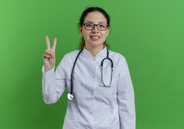 看面带微笑的年轻女医生 穿着医用长袍 戴着听诊器和眼镜 做着和平的标志 隔离在绿色的墙上 留着复印空间眼镜和平微笑