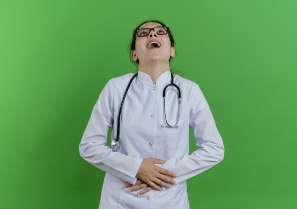女年轻的女医生穿着医用长袍 戴着听诊器和眼镜 抬起头 抱着肚子 笑着 隔离在绿色的墙壁上 还有复印空间医疗长袍穿