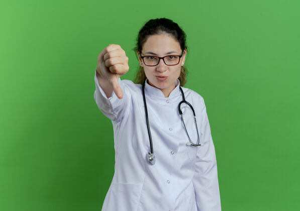 医疗年轻的女医生穿着医用长袍 戴着听诊器 戴着眼镜 拇指朝下 被隔离在绿色的墙上 还有复印空间拇指显示不愉快