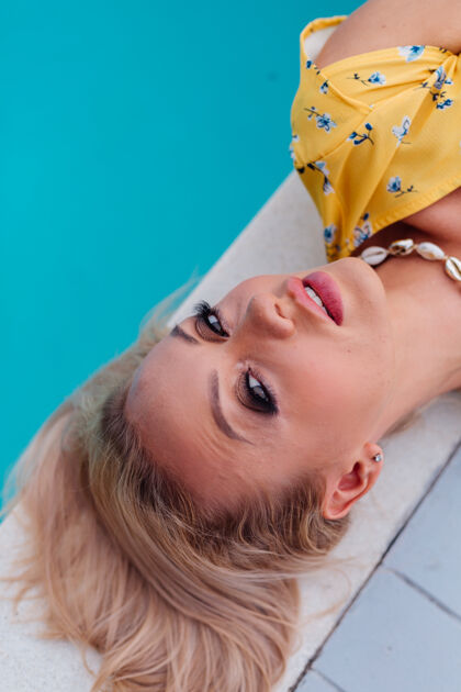 珠宝穿着黄色夏装的放松冷静的女人的肖像躺在蓝色游泳池边 戴着时髦的贝壳项链配件肖像风格