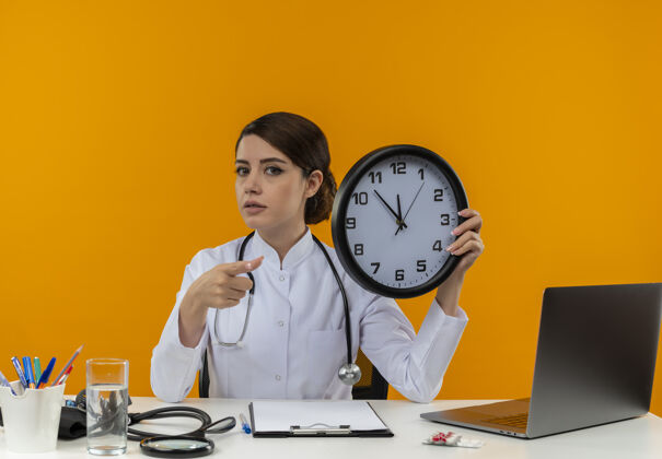 听诊器自信的年轻女医生穿着医用长袍和听诊器坐在桌子旁 带着医疗工具和笔记本电脑 拿着隔离在黄色墙上的时钟长袍拿着工具