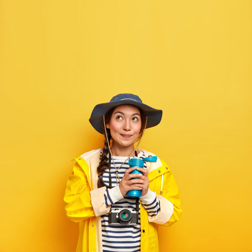 摄影可爱体贴的女孩 戴着时髦的帽子 穿着防护雨衣 拿着热水瓶 拿着复古相机拍照 创造内容女性保温瓶年轻人