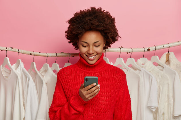 电子卷发开朗的女人 表情欢快 用手机上网支付 穿着红色毛衣 站在衣衫褴褛的背景下 隔离在粉色背景下欢呼编织商店