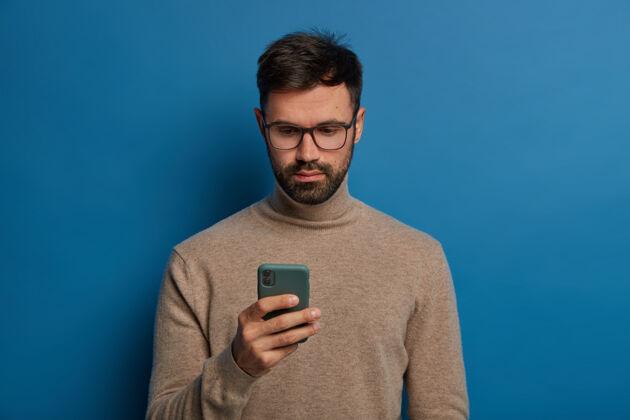 电话严肃的男性使用现代智能手机 专注的注视着蓝色背景上孤立的显示器工作年轻短信
