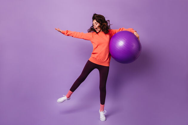 身体身穿黑色紧身裤和橙色连帽衫的活跃女性 在紫色墙上与fitball一起跳跃健身球女性运动服
